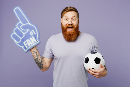 Young fun man fan wears basic t-shirt casual clothes foam 1 fan glove finger up cheer up support football sport team hold in hand soccer ball watch tv live stream isolated on plain purple background.