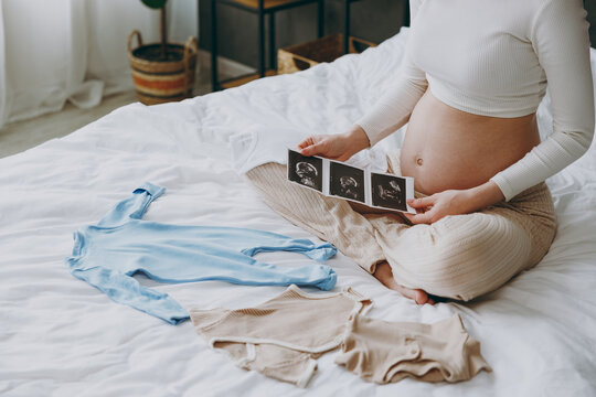 Cropped young pregnant woman in white pyjamas hold in hand little clothes picture of ultrasound test rest relax spend time in bedroom room lounge home. Maternity family pregnancy expectation concept