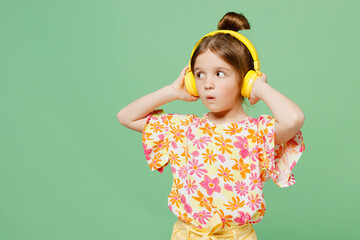 Little surprised child kid girl 6-7 year old wearing casual clothes headphones listen music look aside isolated on plain green background studio portrait. Mother's Day love family lifestyle concept.