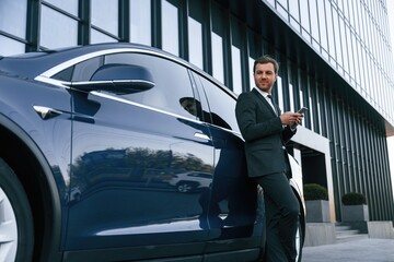 Holding smartphone. Businessman is standing near his car outdoors