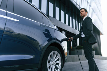 Man in suit and tie is holding charger and standing near his electric car outdoors