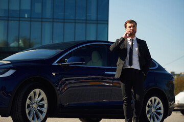 Conversation by phone. Business building behind. Man in suit and tie is standing near his car outdoors