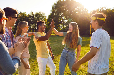 Give me high five. Cheerful company of different young people have fun together during summer walk in park. Dark-skinned guy and Caucasian girl laugh out loud and give each other high five.