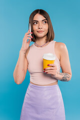 to go coffee, happy and tattooed young woman with short hair and nose piercing holding paper cup and talking on smartphone on blue background, generation z, summer trends, attractive, coffee culture