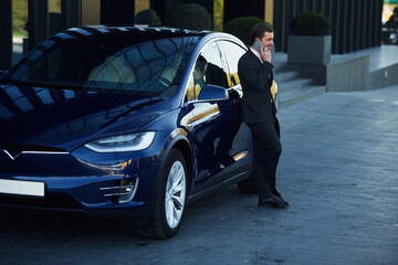 Standing, in black suit. Businessman is near his car outdoors