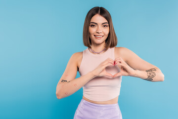 Fototapeta na wymiar positivity and love, happy young woman with short hair, tattoos and nose piercing showing heart gesture with hands on blue background, generation z, cheerful, casual attire