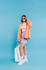 trendy shopping, brunette young woman in tank top, skirt, sunglasses and orange shirt posing with shopping bag on blue background, casual attire, stylish posing, gen z, modern fashion