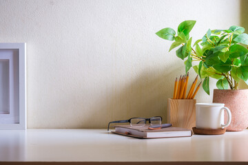 Comfortable workplace with cup of coffee, glasses, picture frame and houseplant on white table.