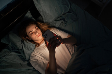 Insomnia, addiction concept. Sleepy exhausted young woman lying in bed using smartphone, top view....