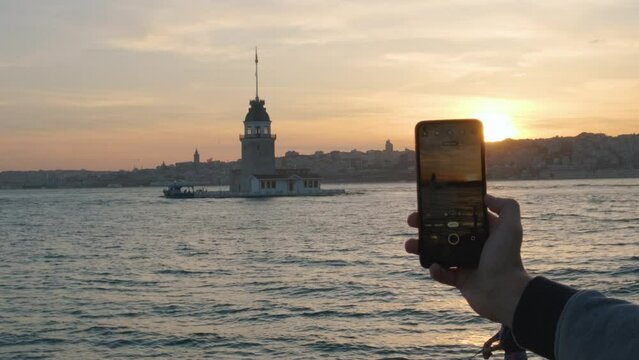 Hand with a phone photographing Maiden's Tower in Bosphorus strait in Istanbul at sunset using camera on his mobile phone. Tourist taking photo on smartphone