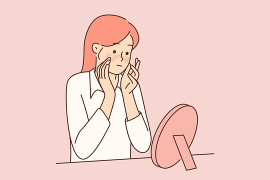 Teenager girl with freckles on face looks in mirror, studying rash or pimples that have appeared