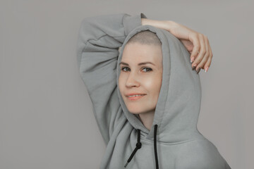 a beautiful young woman with a shaved bald head is dressed in a sweatshirt hood on a light background