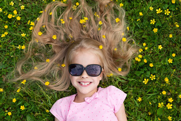 Smiling girl in sunglasses lies on the grass with yellow ranunculus flowers. Happy childhood,...