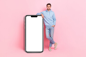 Full length photo of young promoter positive guy wear blue sportswear hoodie stay near big touchscreen menu phone isolated on pink color background