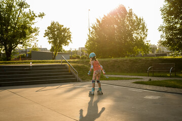 The girl is roller skating at the skate park at sunset. The 8-year-old girl is wearing protective gear and a helmet while roller skating.