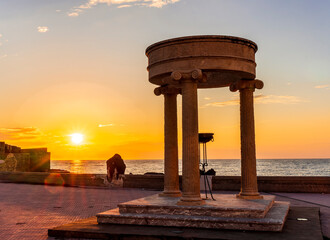 beautiful stone monument with three columns in ancient history greek style with see embarkment and amazing cloudy sunset on background of landscape