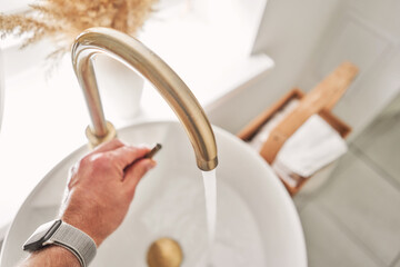 A man's hand touches the water in a beautiful sink with a golden faucet next to an oval mirror and a shelf with hand towels. Close-up of an elegant golden faucet in the bathroom sink next to stylish