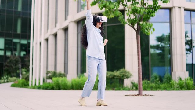 Happy joyful young attractive woman dancing using VR glasses on the street in front of a modern building. Crazy curly brunette female having fun in virtual reality simulator. Dance of a funny teenager