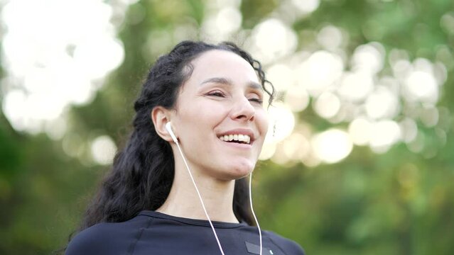Close up. Happy young woman in headphones relaxing with closed eyes standing in urban city park. A smiling fit female brunette breathes deeply and enjoys being in nature outdoors, listening to music
