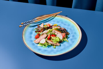 Vivid Asian salad with duck and strawberries, served on a blue ceramic plate with chopsticks. Set against a deep blue backdrop, harsh shadows create a modern, minimalist aestheti