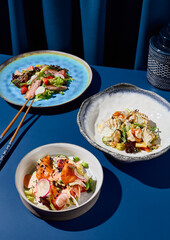 Set of Asian salads presented in a minimalist Asian restaurant interior against a monochromatic blue background with blue curtain. Harsh shadows create a modern still life. Vertical format.