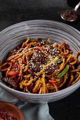 Vertical side view of a stir-fried dish with hand-made wheat noodles, beef, and vegetables, seasoned with a peppery sauce and sesame seeds, displayed on a black background with textiles and spices