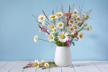 A bouquet of white daisies and other wildflowers in a white vase on a blue background
