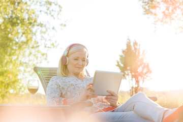 Senior woman headphones wine using digital tablet on lounge chair in sunny backyard