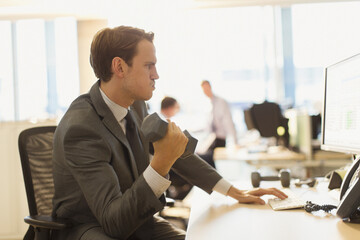 Businessman doing biceps curls with dumbbell at computer in office