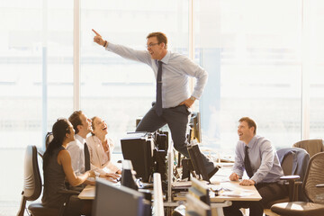 Laughing colleagues watching exuberant businessman dancing on top of desks in office
