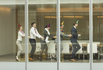 Business people wearing party hats dancing in conga line at conference room window - Powered by Adobe