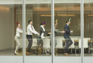 Foto op Canvas Business people wearing party hats dancing in conga line at conference room window © KOTO