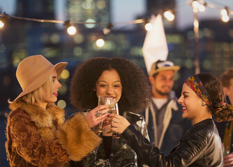 Fototapeta premium Young women toasting champagne glasses at nighttime rooftop party