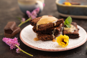 Chocolate brownie with caramel sauce with a cup of coffee on black concrete, side view, selective focus.