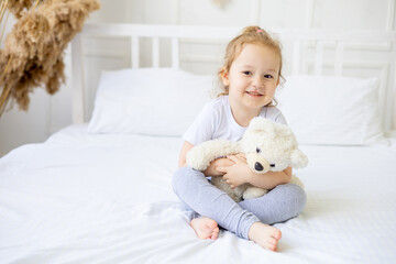 a little cute baby girl on a white cotton bed at home with a soft toy in her hands smiling on the bed at home