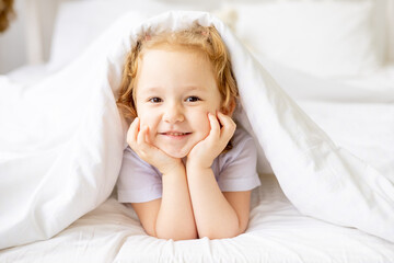 a little cute baby girl on a white cotton bed at home is indulging and having fun smiling on the bed at home with her hands folded under her cheeks