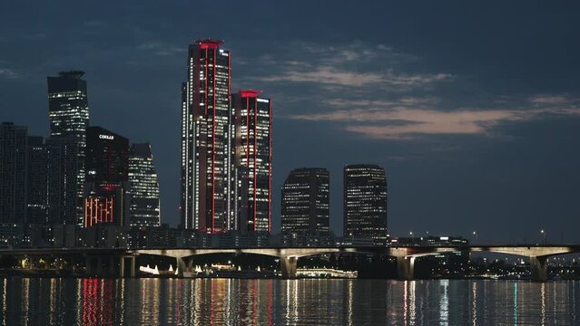 The night time scenery of Yeouido Hangang Riverside in Seoul. Across the Han River on a summer night, you can see a bridge, skyscrapers, and the city lights reflecting on the surface of the river.