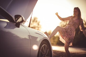 Woman kicking her broken car. Sunset.