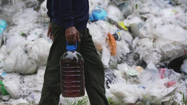 A man carrying a plastic bin filled with biodiesel walks through a large pile of rubbish.