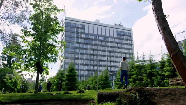 Man in white shirt walks along city park lawn to highrise multi storey building slow motion. Green fir trees decorate side of park