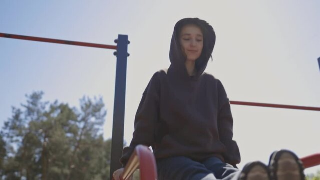 Attractive girl in a sweatshirt on a street sports ground, is engaged in horizontal bars. a caucasian girl is engaged in horizontal bars on a street sports ground. sunny day. shooting from a low angle