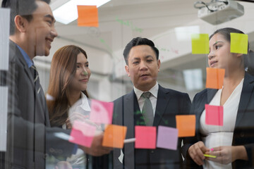 Businessperson teamwork meeting in office and use sticky post notes to brainstorming ideas on glass board. Asian businesswoman planning corporate strategy with colleague for success startup business.