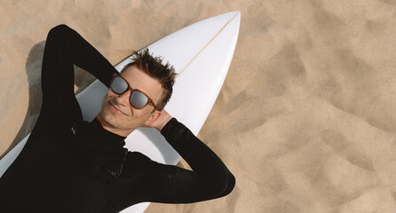 Surfer in wetsuit and sunglasses with surfboard relaxing at beach, sand background. View from above and copy space for text. 