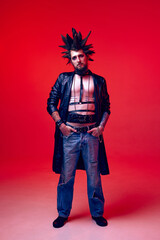 Portrait of young guy in image of punk with extraordinary hairstyle, makeup posing against red...
