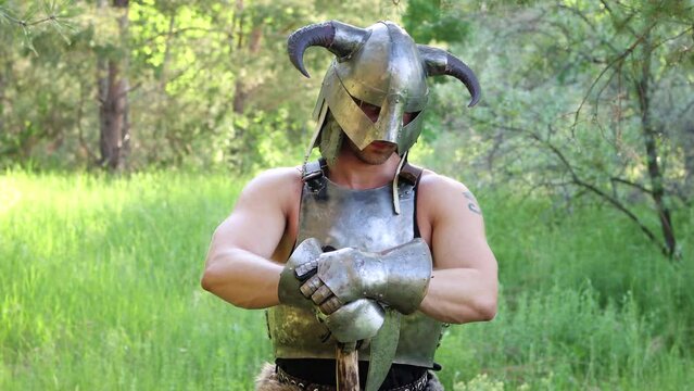 Smooth camera movement, selective focus. Portrait of a warrior in a horned helmet, a steel breastplate with a two-handed ax in his hands, posing against the backdrop of the forest. 