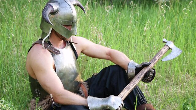 Smooth camera movement, selective focus. Portrait of a warrior in a horned helmet, a steel breastplate with a two-handed ax in his hands, sitting in tall grass against the backdrop of the forest.
