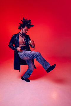 Emotional young man punk in extraordinary clothes, with makeup playing illusion guitar against red studio background in neon light. Concept of music, lifestyle, subculture, art, youth, human emotions