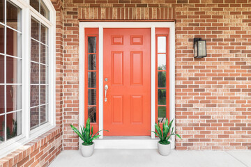 A home's red front door surrounded by red brick and plants sitting in front of the windows. - obrazy, fototapety, plakaty
