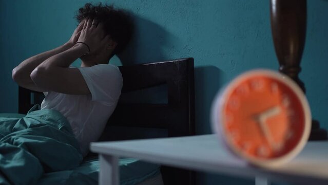 Unhappy young man with insomnia sitting on bed next to alarm clock at night, suffering headache. Insomnia