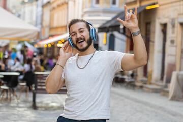 Happy relaxed overjoyed bearded young man in wireless headphones choosing, listening favorite energetic disco rock n roll music dancing outdoors. Guy walking in urban sunshine crowded city street